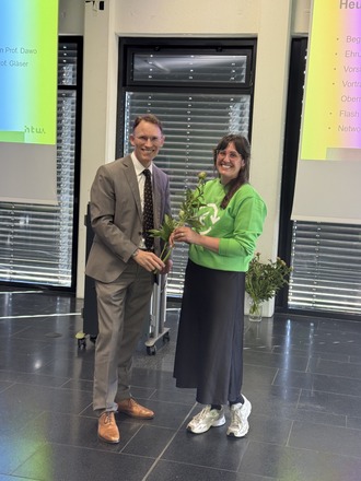 Prof. Dr. Sascha Dawo überreicht einer Absolventin in einem grünen HTW-Hoodie eine Rose vor einer Präsentationsleinwand.