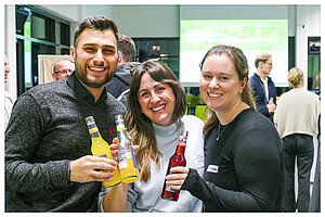 Drei junge Alumni stehen eng beieinander in einem modern beleuchteten Veranstaltungsraum der HTW Berlin und lächeln freudig in die Kamera.